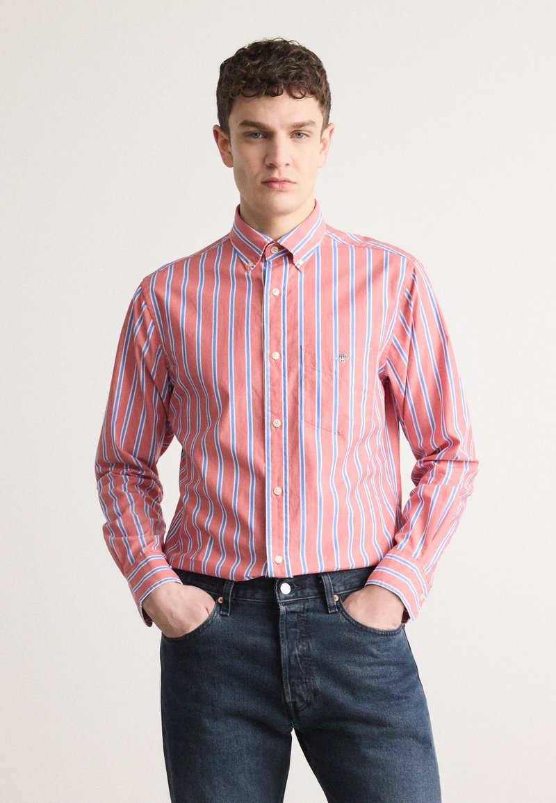 Camisa de botones a rayas rojas y azules con cuello puntiagudo y un pequeño bolsillo en el pecho, combinada con jeans azul oscuro. Textura de tela suave.