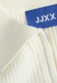 Hladký krémový svetr s užším límcem na zip. Má modrou štítek s nápisem "JJXX." Měkká textura a výrazné svislé pruhy na látce.