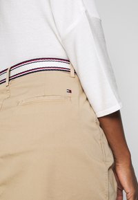 Pantalones beige con una textura suave, que presentan una cinturilla a rayas en blanco, rojo y negro. Incluyen un bolsillo trasero con un pequeño logo.