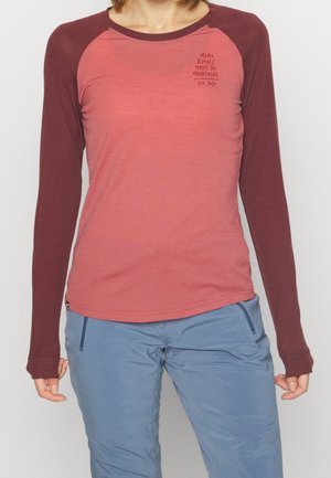 Femme portant un t-shirt bicolore manches longues rouge et bordeaux avec un texte sur la poitrine, associé à un pantalon bleu clair avec des poches zippées.