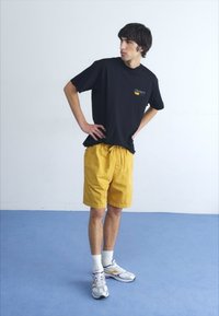 T-shirt nera in cotone con logo, pantaloni corti gialli, calzini bianchi e scarpe da ginnastica argentate. Posizione in piedi su un pavimento blu con sfondo bianco.