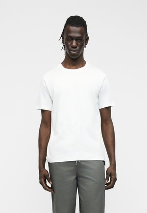 Homme debout face à l'avant, portant une chemise blanche unie à manches courtes et un pantalon gris, sur un fond clair uni.