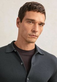 Dunkelgraues Strick-Poloshirt mit Kragen und zwei Knöpfen. Wird über einem dunkelbraunen Rundhalshemd getragen. Glatte Textur, eng anliegendes Design.