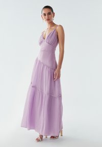 Maxikleid in Lavendel mit Spaghettiträgern, strukturierter Stoff, Empire-Taille und mehrlagigem Design. Weiße Sandaletten mit Absatz betonen den Look.