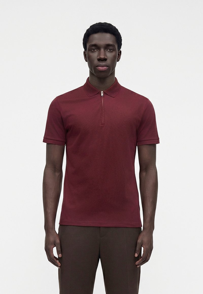 Homme portant un polo bordeaux à manches courtes avec une fermeture éclair sur le devant et un pantalon marron foncé, debout devant un fond clair uni.