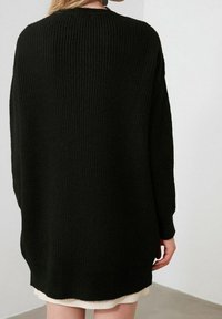 Personne portant un pull en tricot noir ample par-dessus une jupe blanche, debout à l'intérieur contre un mur uni.
