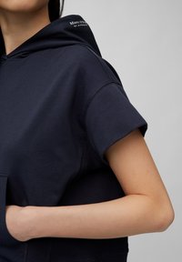 Sudadera azul marino confeccionada en una mezcla de algodón, con mangas cortas, bolsillo frontal y detalle del logo en la capucha. Textura suave y lisa.