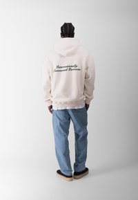 Sudadera crema con texto bordado en verde en la parte de atrás que dice "Ropa Reconocida Internacionalmente," combinada con pantalones azul claro y zapatos negros.