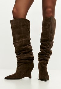 Barna velúr térdig érő csizma, laza kialakítással és hegyes ék sarkú sarokkal. Texturált anyag puha redőkkel és látható varrásokkal.