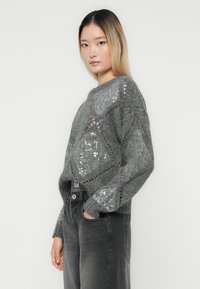 Tânără cu păr drept ombre, purtând un pulover tricotat gri cu modele de diamante cu paiete și blugi negri cu talie înaltă.