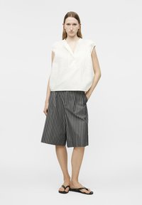 Kvinne iført ermeløs hvit bluse, knelang shorts med svarte og hvite vertikale striper, og svarte flip-flops, stående med en hånd i lommen.