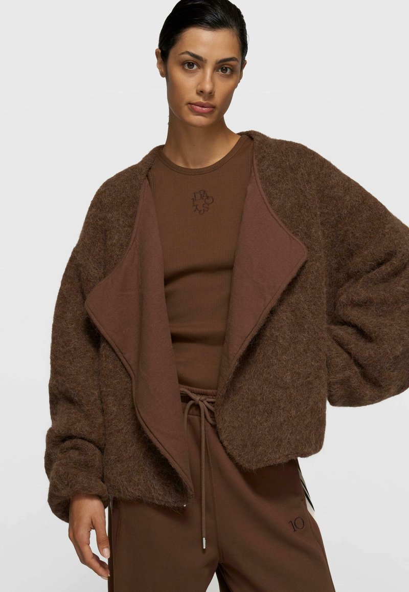 Brauner, flauschiger Cardigan mit drapiertem Vorderteil und weiten Ärmeln, getragen über einem passenden braunen Oberteil. Das Outfit zeichnet sich durch glatte Texturen und subtile Details aus.