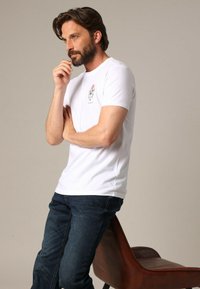 T-shirt blanc à manches courtes avec un petit motif sur la poitrine, associé à un jean bleu foncé. Le modèle est debout et pensif.