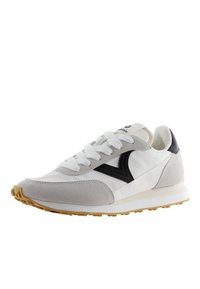 Zapatilla deportiva blanca y gris con materiales de ante y sintético, detalles en negro, suela de goma texturizada y cordones blancos. Estilo inspirado en lo retro.