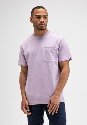 Junger Mann steht, trägt ein hellviolettes kurzärmeliges T-Shirt mit Brusttasche und dunkle Hose, schaut zur Seite.