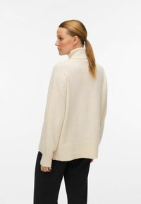 Kremna turtleneck pulover s sproščenim krojem, rebrastimi manšetami in robom, z mehko teksturo. Uporabljeno z temnimi hlačami, pogled prikazuje zadnji profil.