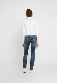 Top a maniche lunghe bianco aderente con collo alto abbinato a jeans in denim blu con una leggera sbiaditura e tasche posteriori con cuciture decorative.