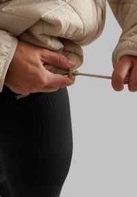 Beige quilted jacket with a pull cord and toggle detail, worn over black ribbed leggings, showing hands adjusting the jacket's fit.