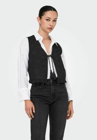Zwarte geborduurde vest met een striksluiting en franjes, over een wit overhemd met knopen gedragen en gecombineerd met zwarte jeans.