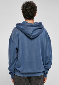 Felpa blu con tessuto testurizzato, vestibilità oversize e cappuccio con coulisse. Presenta polsini e orlo a coste. La vista posteriore mostra i dettagli della cucitura.