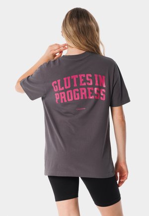 Kobieta z długimi włosami, ubrana w szarą koszulkę z różowym napisem „GLUTES IN PROGRESS” oraz czarne spodenki rowerowe, zwrócona tyłem, z ręką przy szyi.