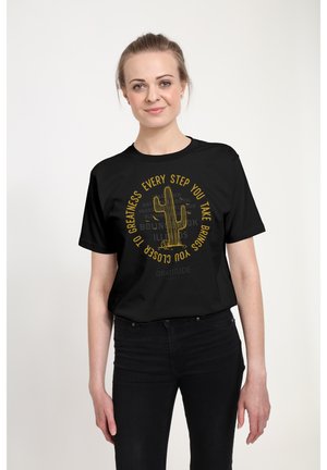 Ung kvinde iført sorte jeans og en sort T-shirt med en gul kaktus og motiverende tekst, smilende foran en ensfarvet hvid baggrund.