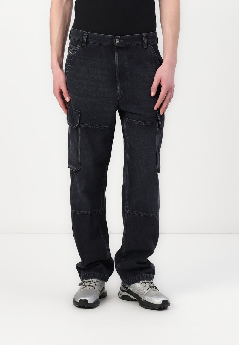 Personne portant un pantalon cargo noir et des chaussures de sport argentées, debout devant un fond blanc uni.