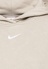 Sweat à capuche beige avec un logo Nike swoosh blanc sur la poitrine et des coutures visibles le long du bord de la capuche.