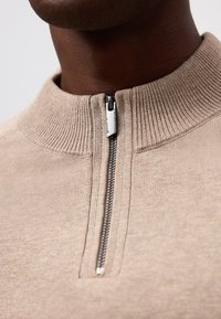 Maglione beige con zip frontale e colletto alto. Presenta una texture a coste e una zip metallica. Vista ravvicinata che si concentra sul collo e i dettagli della zip.