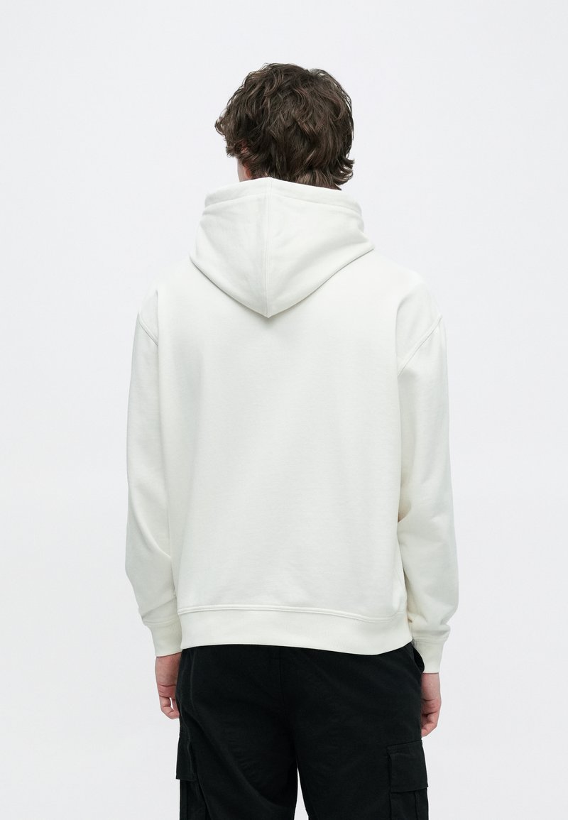 Felpa bianca con cappuccio con coulisse, tasca a marsupio e polsini a costine. Tessuto morbido con texture liscia. Vista posteriore che mostra i dettagli delle cuciture.