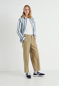 Gestreiftes blau-weißes Hemd mit Knöpfen über einem weißen T-Shirt, khakifarbene, gefaltete Hose und marineblaue Sneaker mit roten und weißen Akzenten.