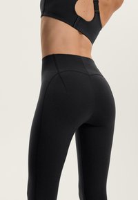 Kobieta w czarnych legginsach o wysokim stanie i czarnym staniku sportowym, stojąca z uniesionymi ramionami na jasnym, jednolitym tle.