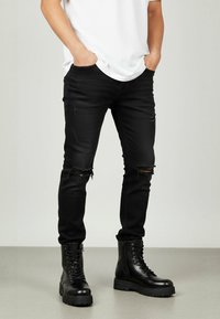 Jeans negri din denim deteriorat cu găuri la genunchi, asortați cu bocanci negri cu șireturi. Modelul poartă un tricou alb.