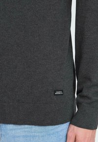 Suéter de punto gris oscuro con un dobladillo y puños acanalados. Presenta una pequeña etiqueta negra cerca del dobladillo con texto blanco, que se muestra sobre un fondo de mezclilla clara.