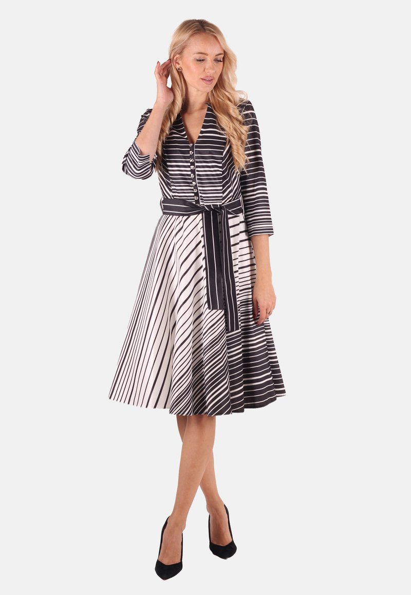Schwarz-weiß gestreftes Kleid mit V-Ausschnitt, langen Ärmeln, durchgehender Knopfleiste und taillierter Taille; ausgestellter Rock mit unterschiedlich breiten Streifen.