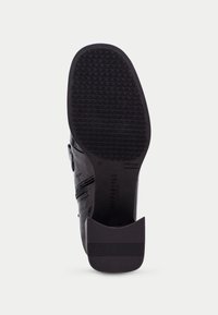 Suela de zapato negra con un fondo de goma texturizada, tacón ovalado y un nombre de marca en relieve, junto con un delantero de cuero suave y detalle de cremallera.