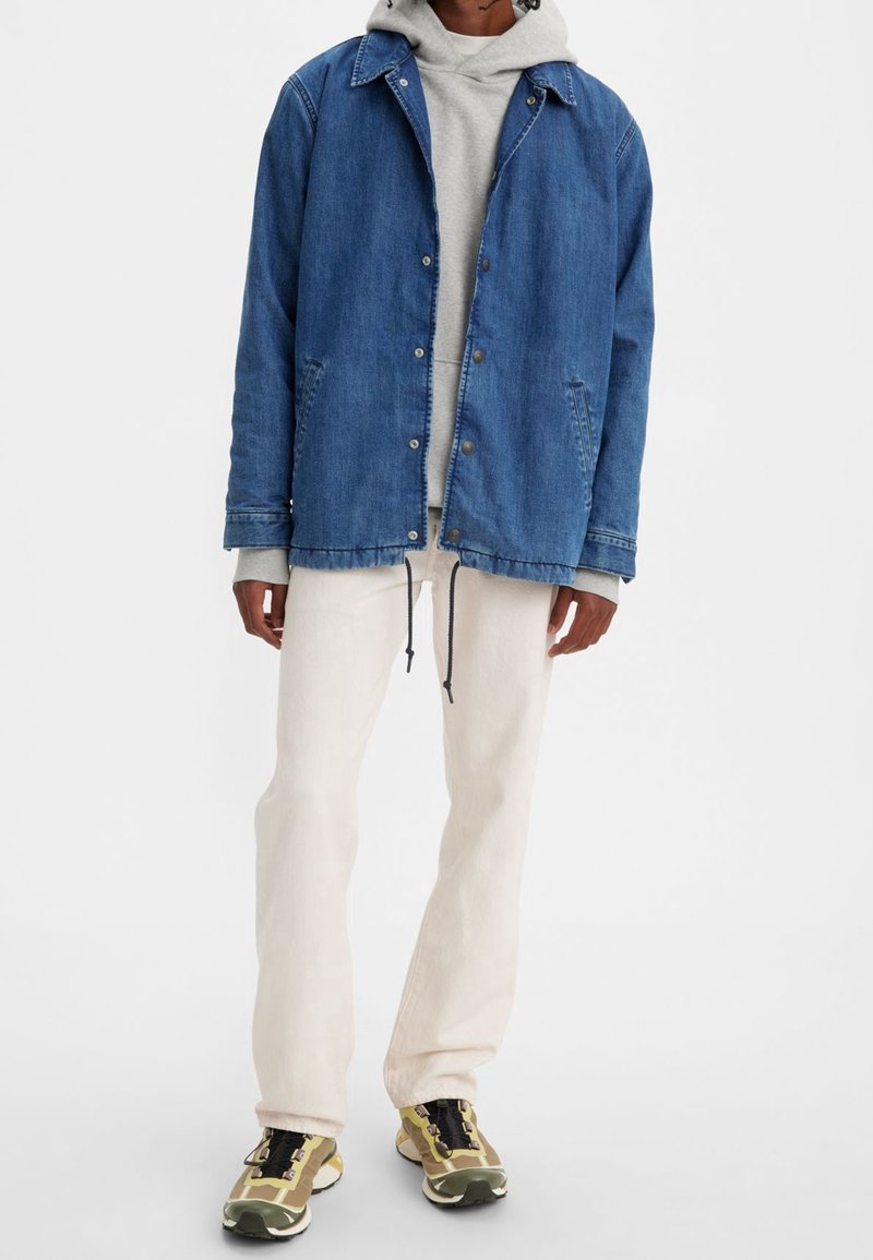 Chaqueta de denim, azul, con botones de presión, dos bolsillos laterales, superpuesta a una sudadera gris; combinada con pantalones de color claro y zapatillas con acentos amarillos.