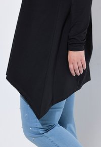 Czarna tunika z linią dołu opadającą na bokach, zestawiona z jasnoniebieskimi dżinsami ozdobionymi perłami wzdłuż zewnętrznych szwów.