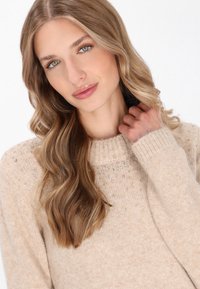 Maglione beige lavorato a maglia con scollatura a coste, rifinito con piccoli dettagli argentati sulle spalle, caratterizzato da una finitura morbida e strutturata.