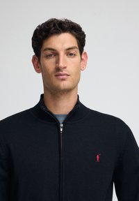 Svart stickad zip-up tröja med ribbad krage och mudd, med en liten röd broderad logotyp på vänster bröst. Enkel texturerad tyg.