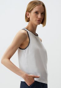 Witte mouwloze top met een gestructureerde afwerking en zwarte zigzagafwerking langs de halslijn. Heeft een ontspannen pasvorm en zijzakdetails.