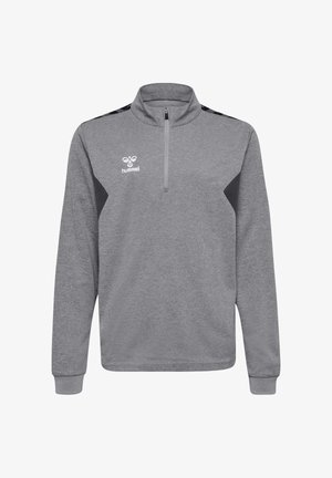 Junge steht mit verschränkten Armen, trägt einen grauen Pullover mit Viertelreißverschluss, weiße Shorts, weiße Socken und schwarze Turnschuhe vor einfachem Hintergrund.