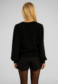 Maglione nero con maniche a sbuffo testurizzate, polsini e orlo a coste. Tessuto del corpo liscio. Indossato con pantaloni neri, capelli parzialmente visibili.