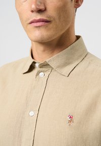Cămașă din in bej cu guler clasic, având butoane albe și un accent brodat mic al unei figuri pe partea stângă.