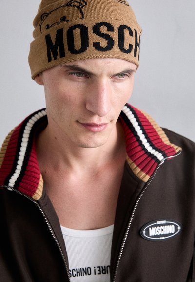 Pruun tõmblukk-jakk rööbastega kraega punases, mustas ja beežis. Pruun kootud beanie musta "MOSCHINO" logoga. Valge särk all.