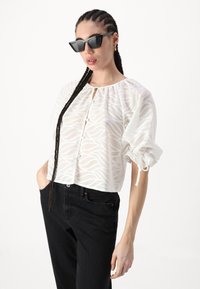 Bluză albă croppată, cu un model texturat în valuri, având un decolteu rotund, închidere cu buton și mâneci bufante. Asortată cu blugi negri.