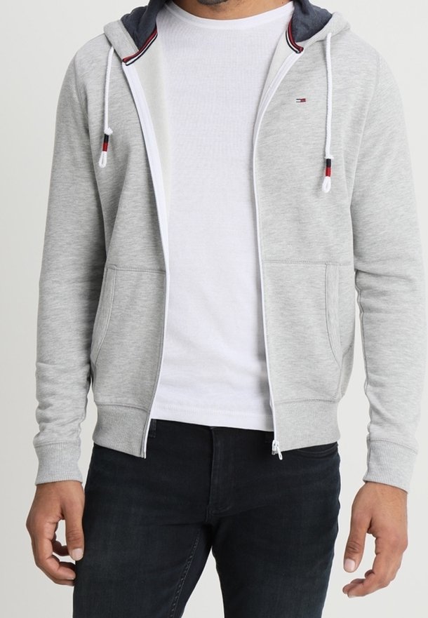 Homme portant un sweat à capuche gris clair zippé avec cordons, par-dessus un t-shirt blanc et un jean foncé, debout devant un fond uni.