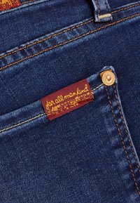 Farkkutakissa on tummansininen väri oranssilla ompelella. Punainen etiketti keltaisella tekstillä on ommeltu taskuun, jossa on bränditietoja.