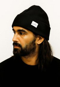 Black ribbed beanie with a white fabric label on the side. The person has long hair and a beard, wearing a black shirt.