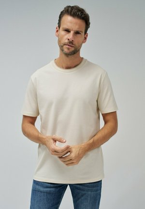 Camiseta beige de manga corta con cuello redondo, tejido texturizado y un sutil logo en el pecho, combinada con jeans azules.